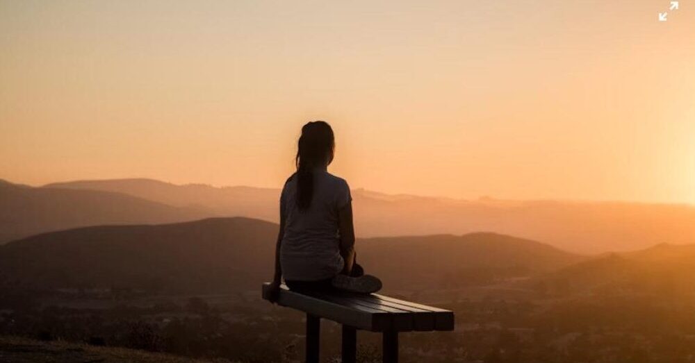 Woman sitting on a bench at sunset overlooking mountains, representing mindfulness and slowing down